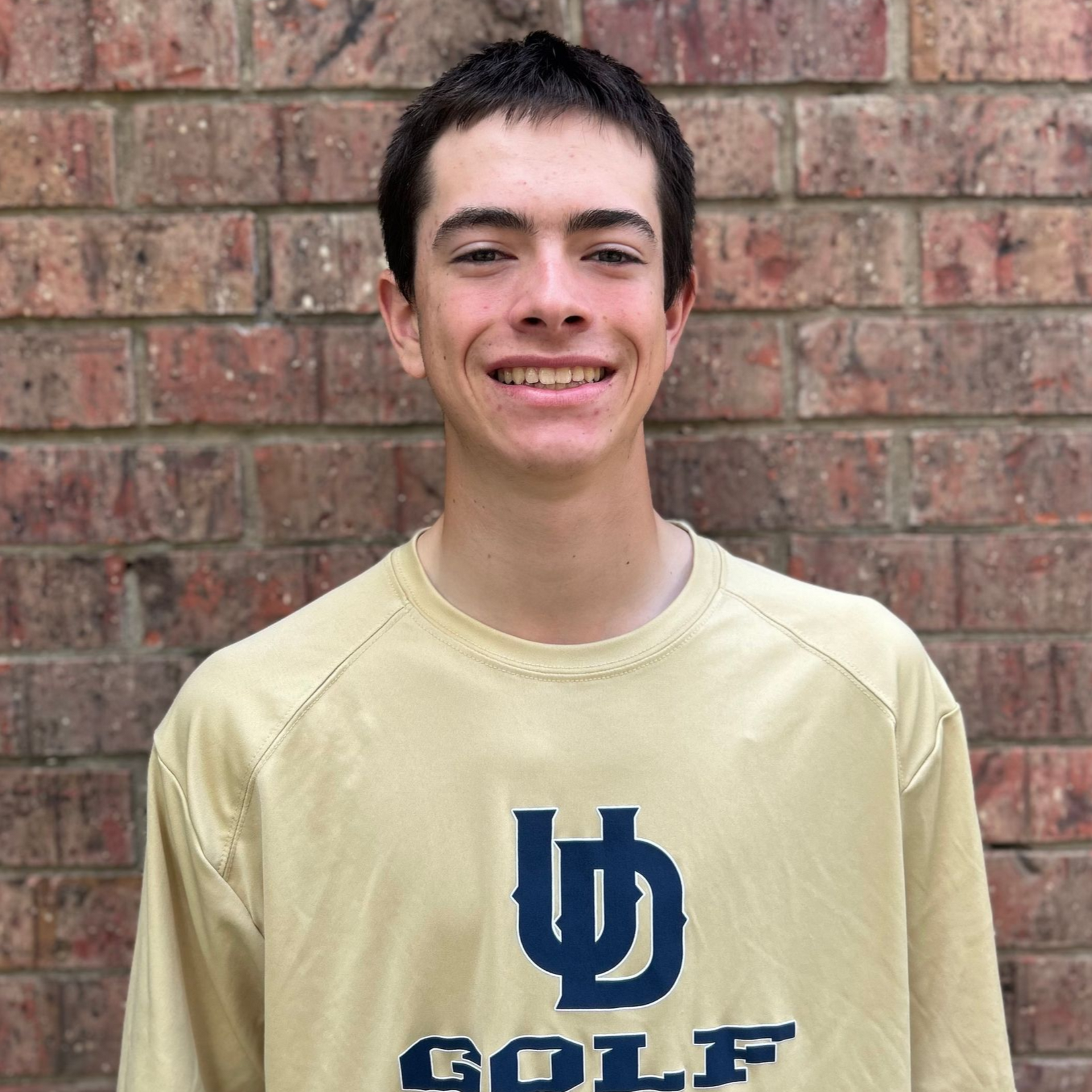 Smiling person in a beige UD Golf shirt standing in front of a brick wall