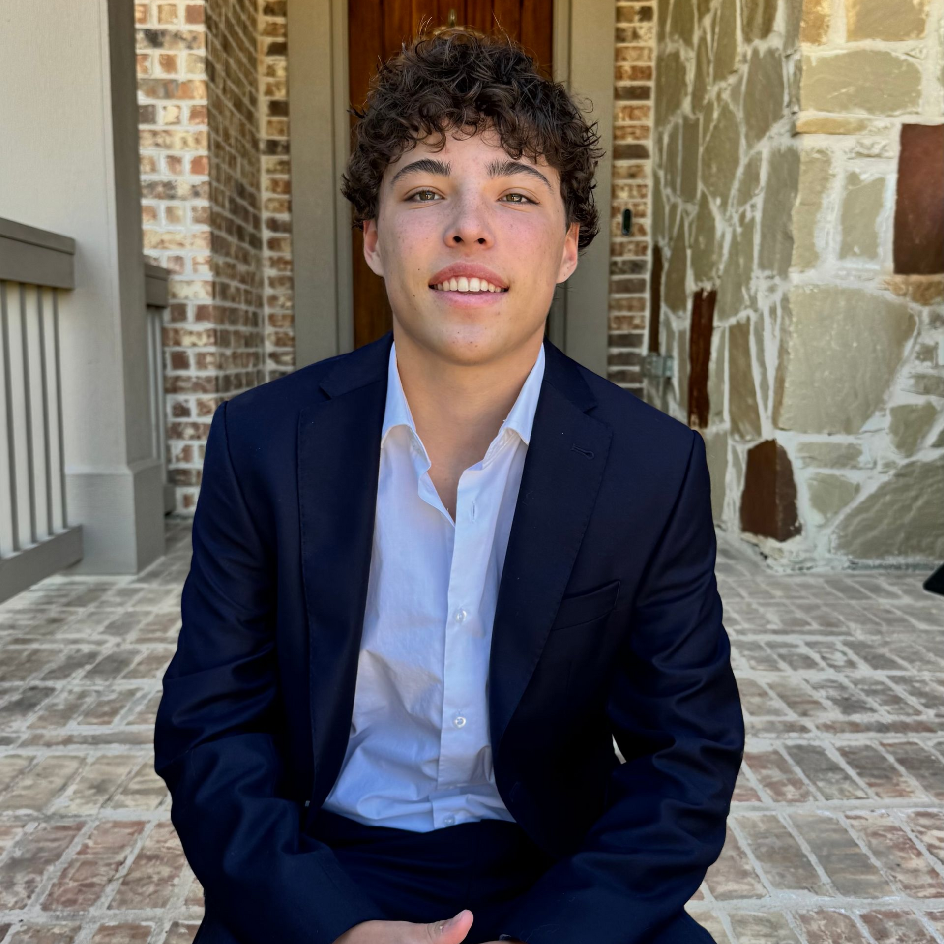 Young man in a navy blazer and white shirt sitting on a stone patio against a brick wall.