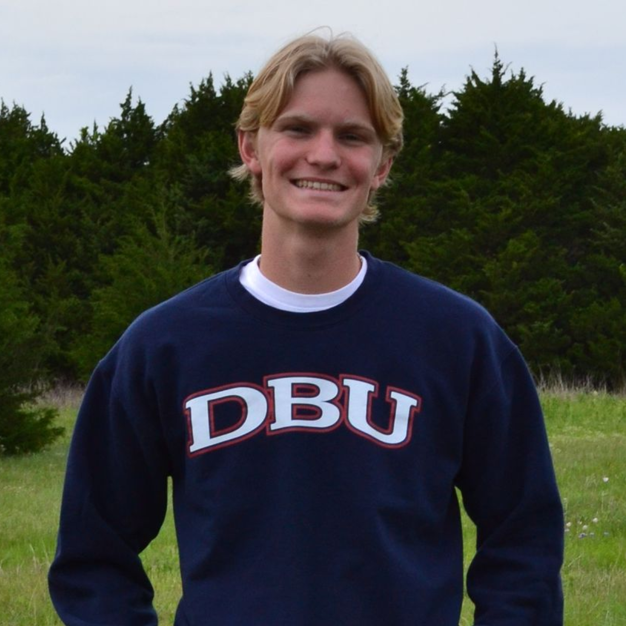 Smiling person in a navy DBU sweatshirt standing outdoors in front of trees