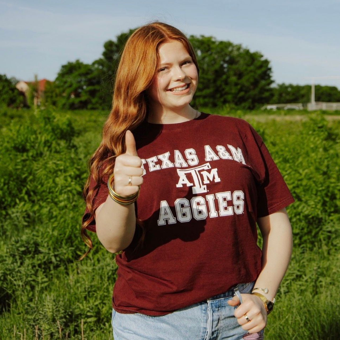 Smiling person in a maroon Texas A&M Aggies shirt giving a thumbs-up in a green field.