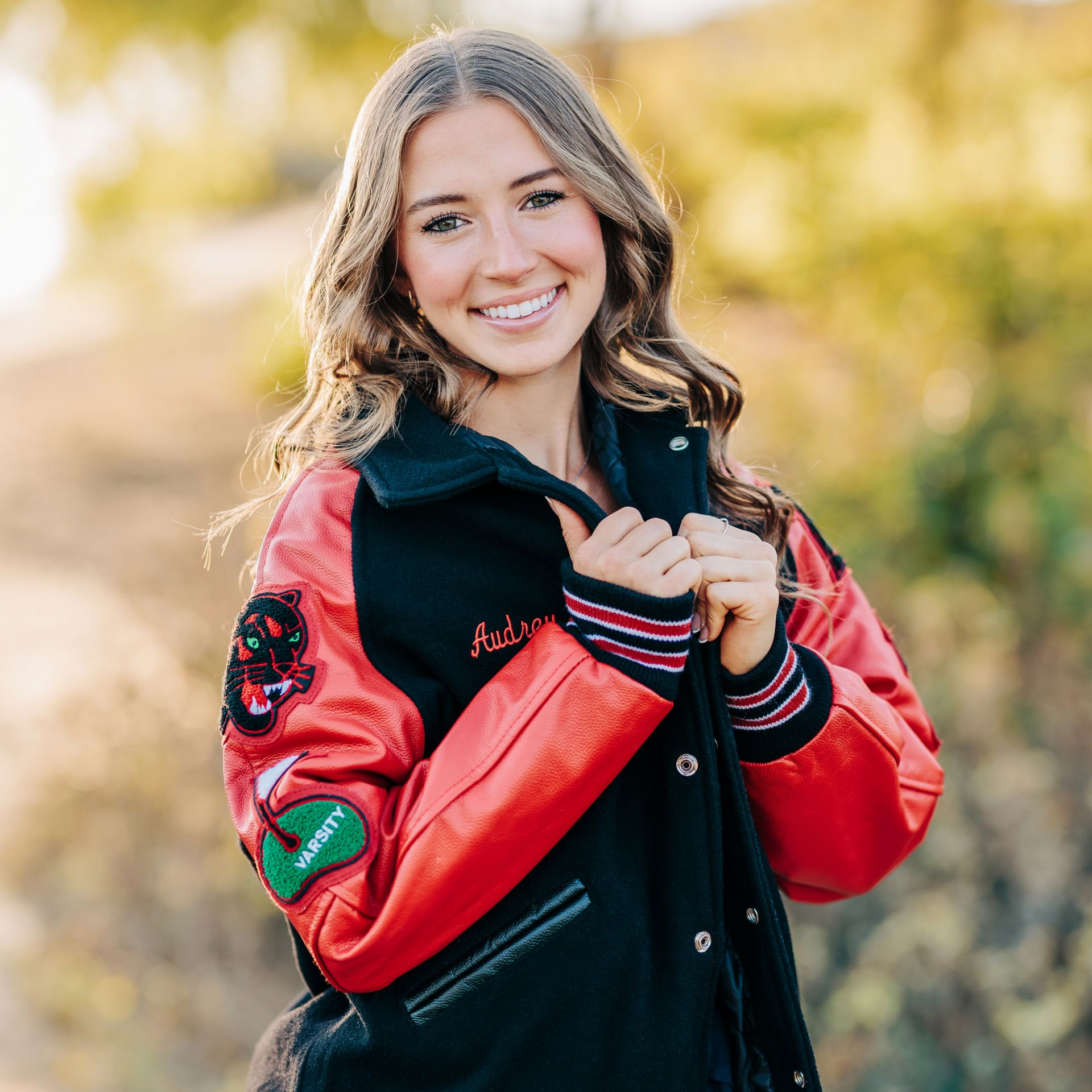 Smiling person in a red-and-black varsity jacket outdoors in soft golden light