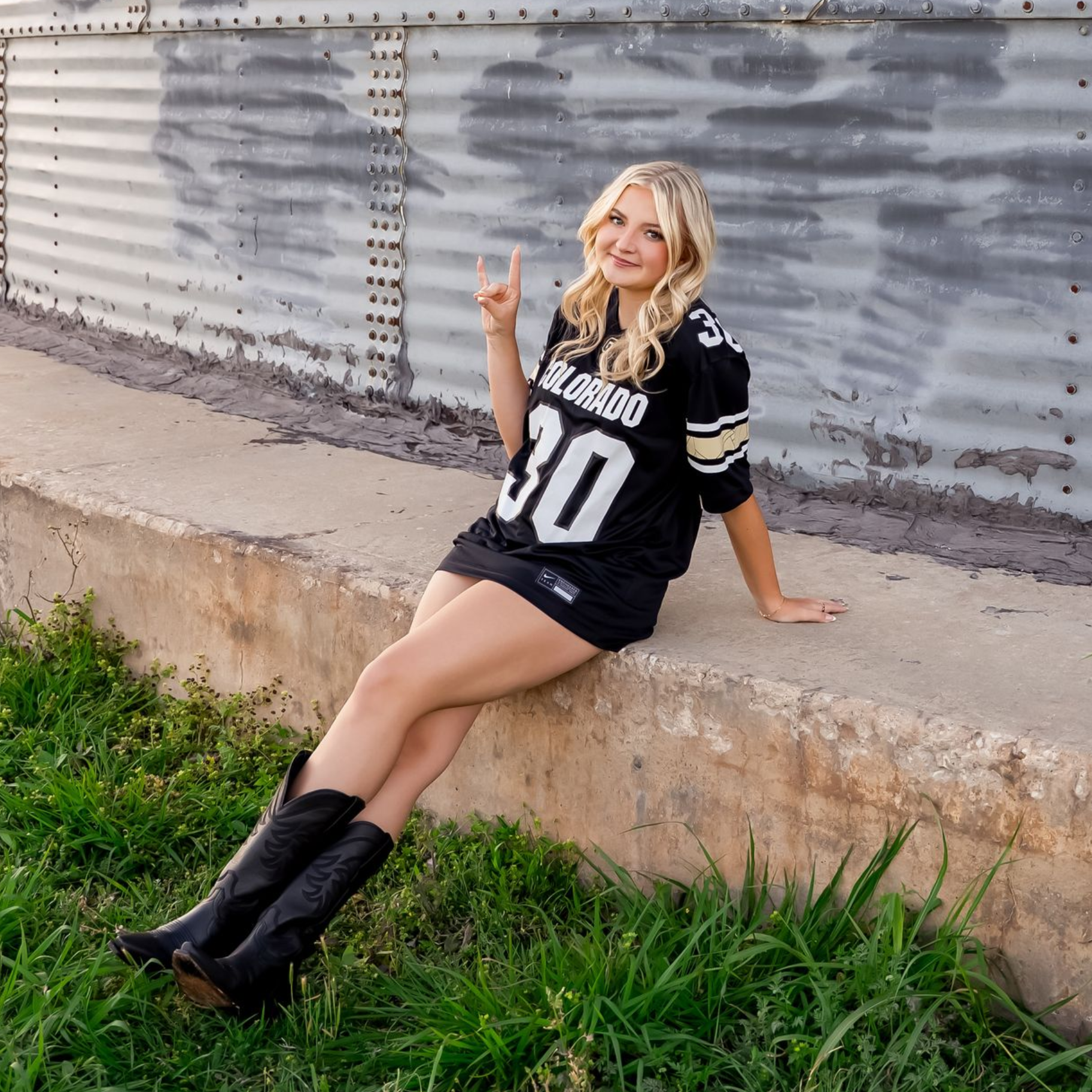 Woman in a black jersey sits on a ledge, smiling and holding up a peace sign by a corrugated metal wall.