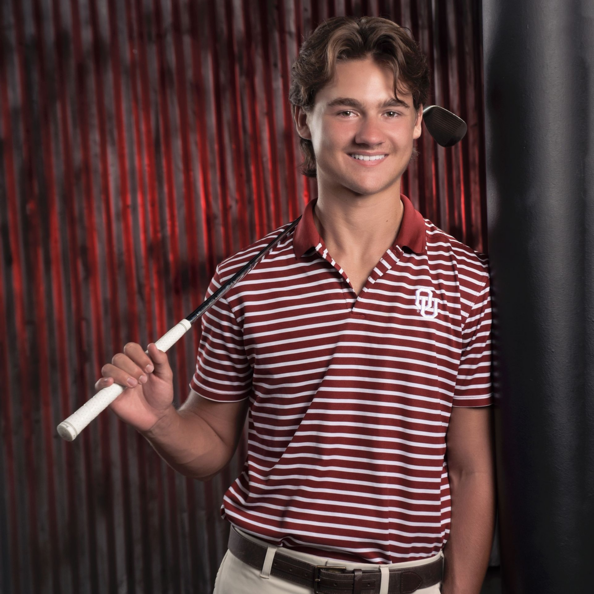 Smiling golfer holding a white club, wearing a maroon striped polo against a dark red and black backdrop