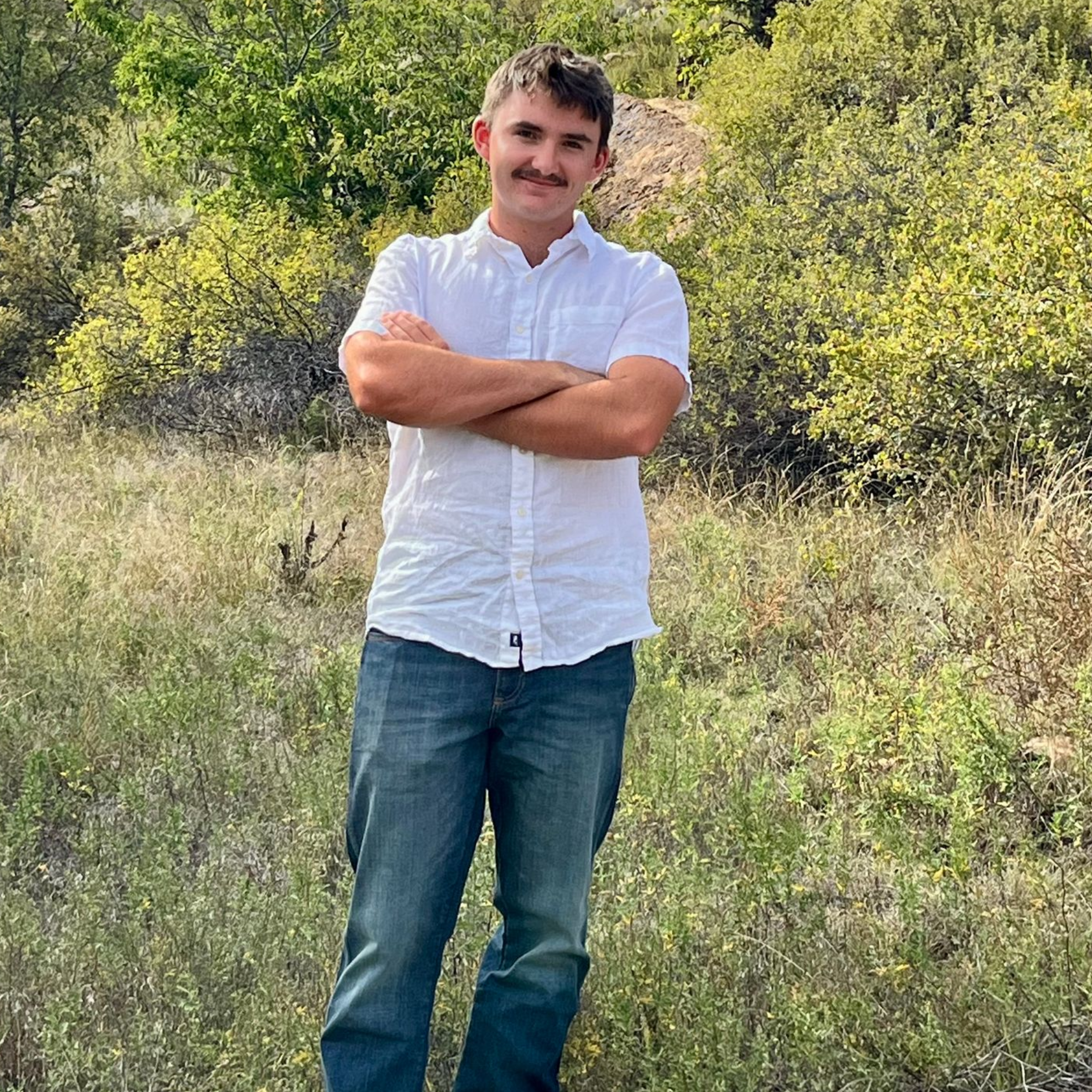 Man standing in a grassy field with arms crossed, wearing a white shirt and jeans, with trees behind him