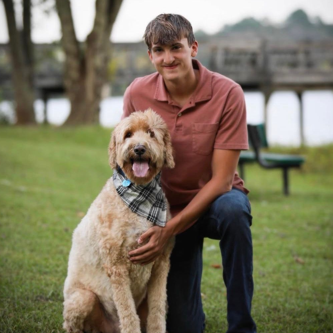 Man kneels beside a tan dog wearing a plaid bandana in a grassy park near water.