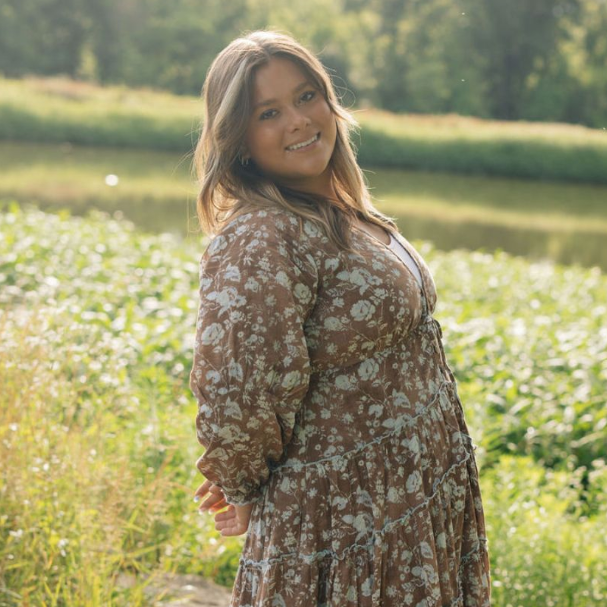 Smiling person in a floral dress standing in a sunlit field near water