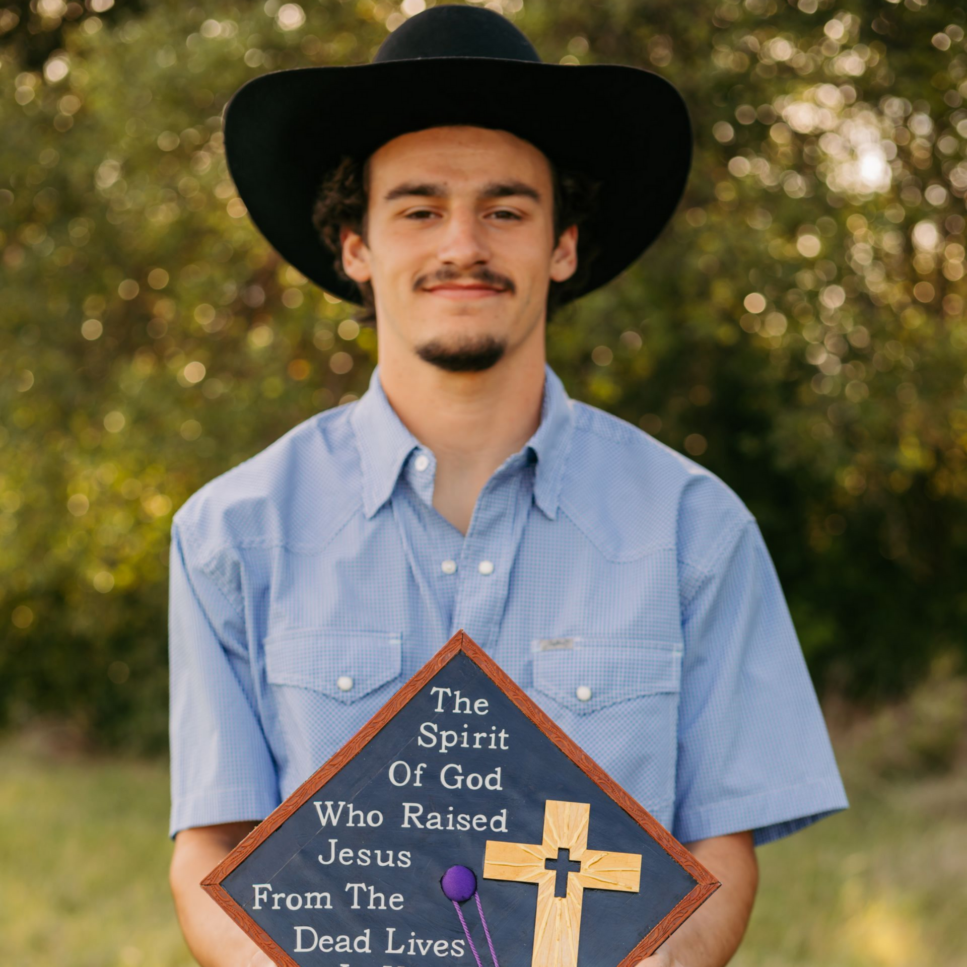 Young man in a black cowboy hat holding a wooden plaque with a religious quote outdoors