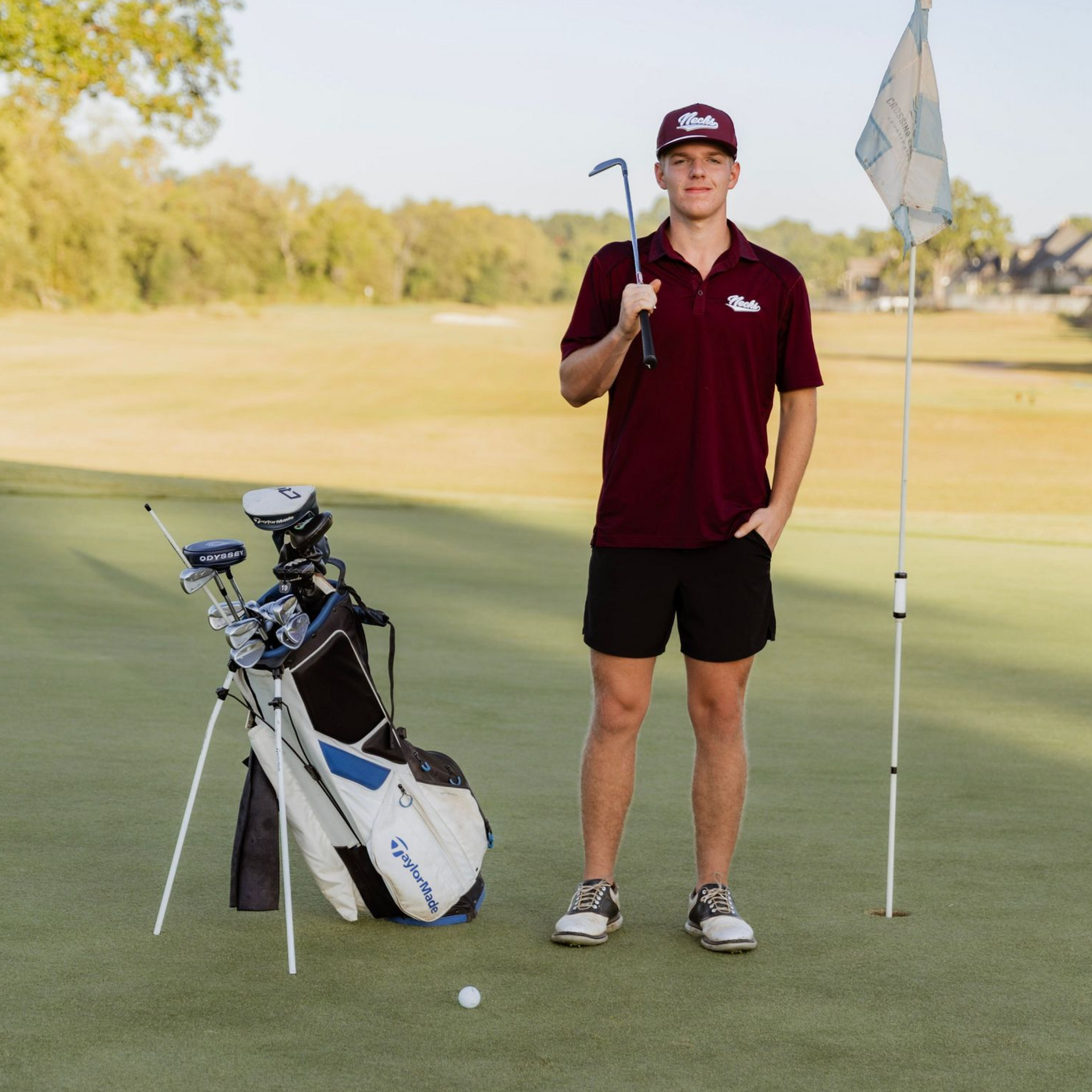 Golfer in maroon shirt standing by golf bag and flag on a course, holding a club