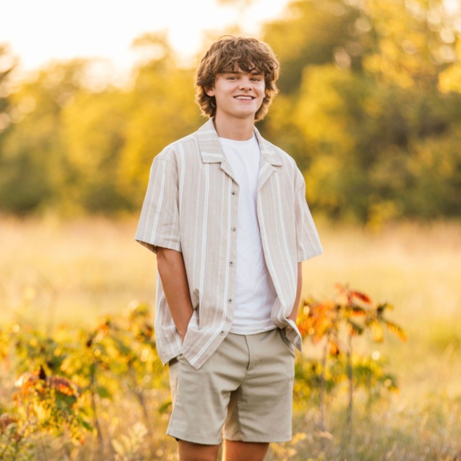 Young person smiling in a sunlit field, wearing a striped shirt, white tee, and beige shorts