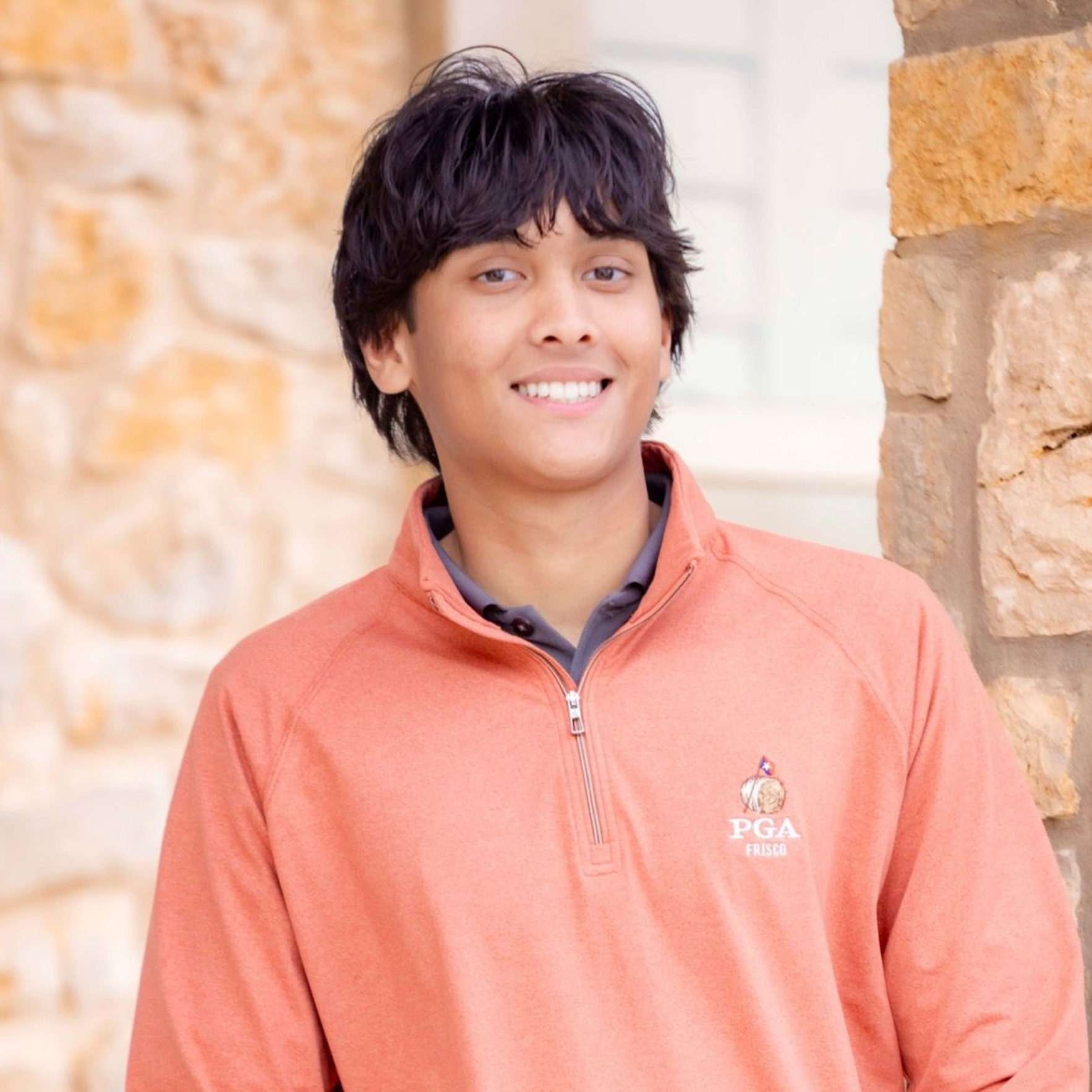 Person smiling in a coral quarter-zip sweater, standing by a stone wall.