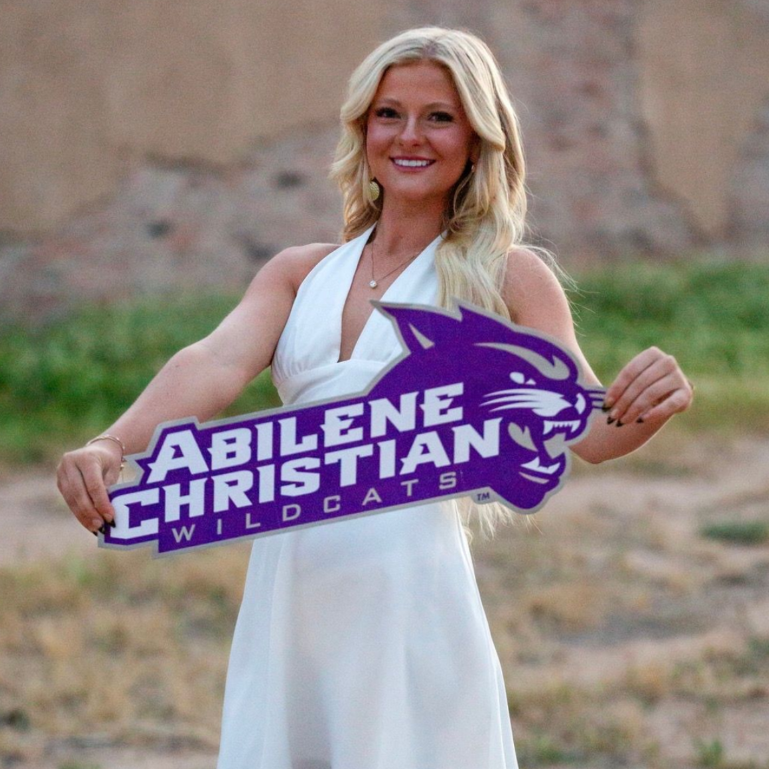 Woman in white dress holding Abilene Christian Wildcats sign outdoors