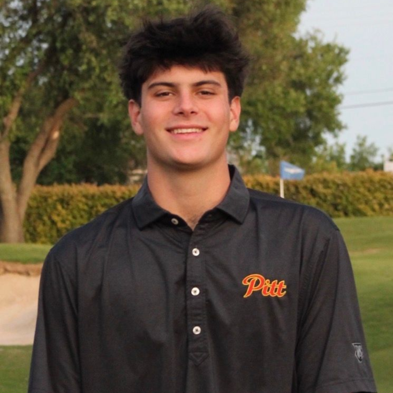 Smiling person in a black Pitt polo shirt standing outdoors with trees in the background