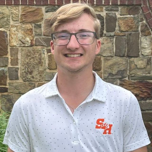 Smiling person in glasses wearing a white polo, standing against a stone wall.