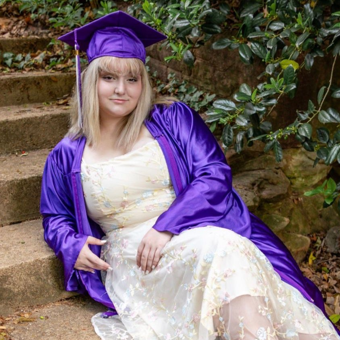 Person in a purple graduation cap and gown sitting on stone steps outdoors.