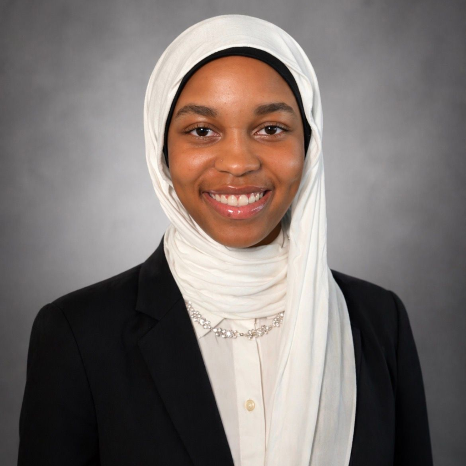 Smiling person in a white hijab and black blazer against a gray studio background