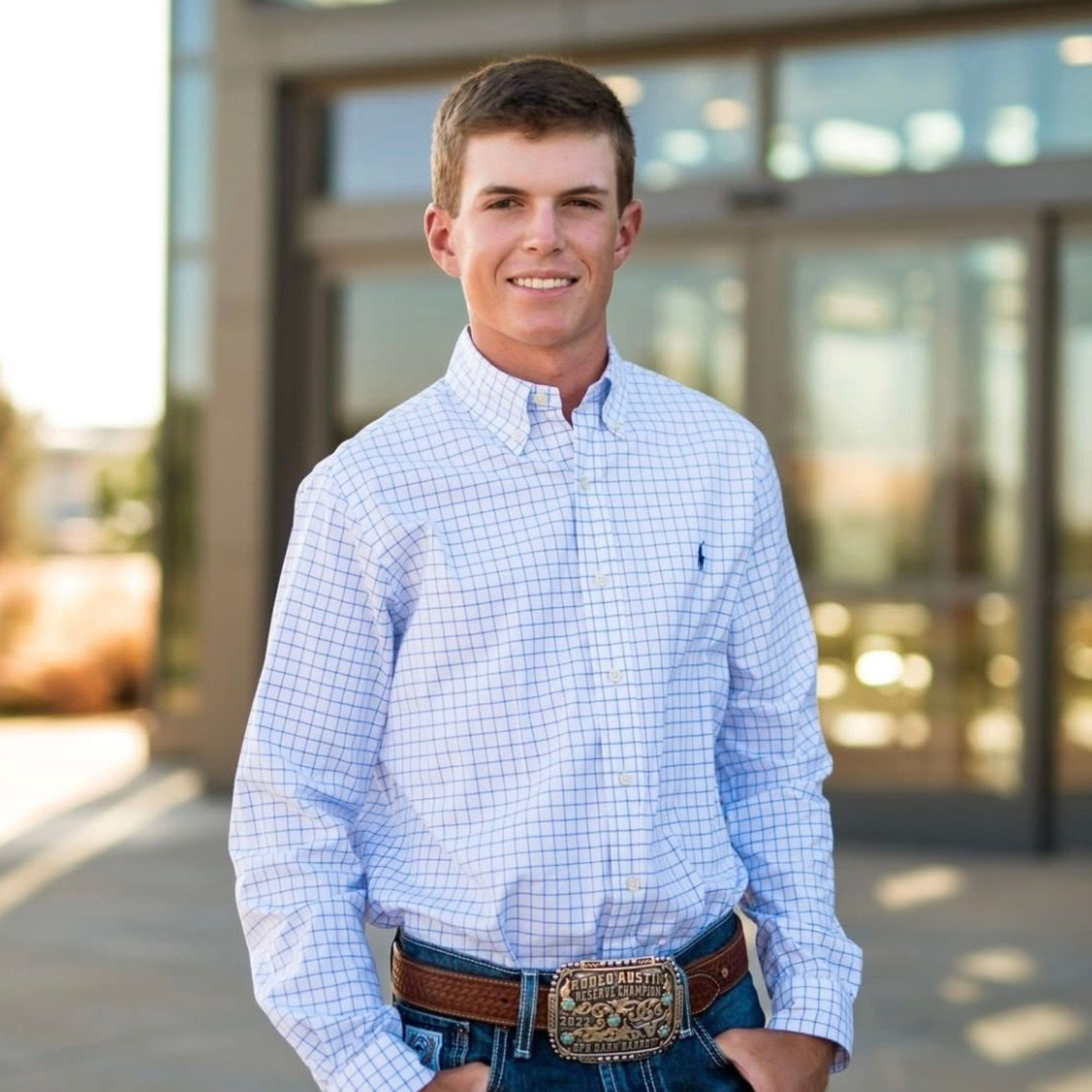 Man in a light blue checkered shirt standing outdoors in front of a building