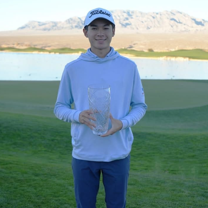 Person holding a clear trophy on a golf course with mountains in the background.