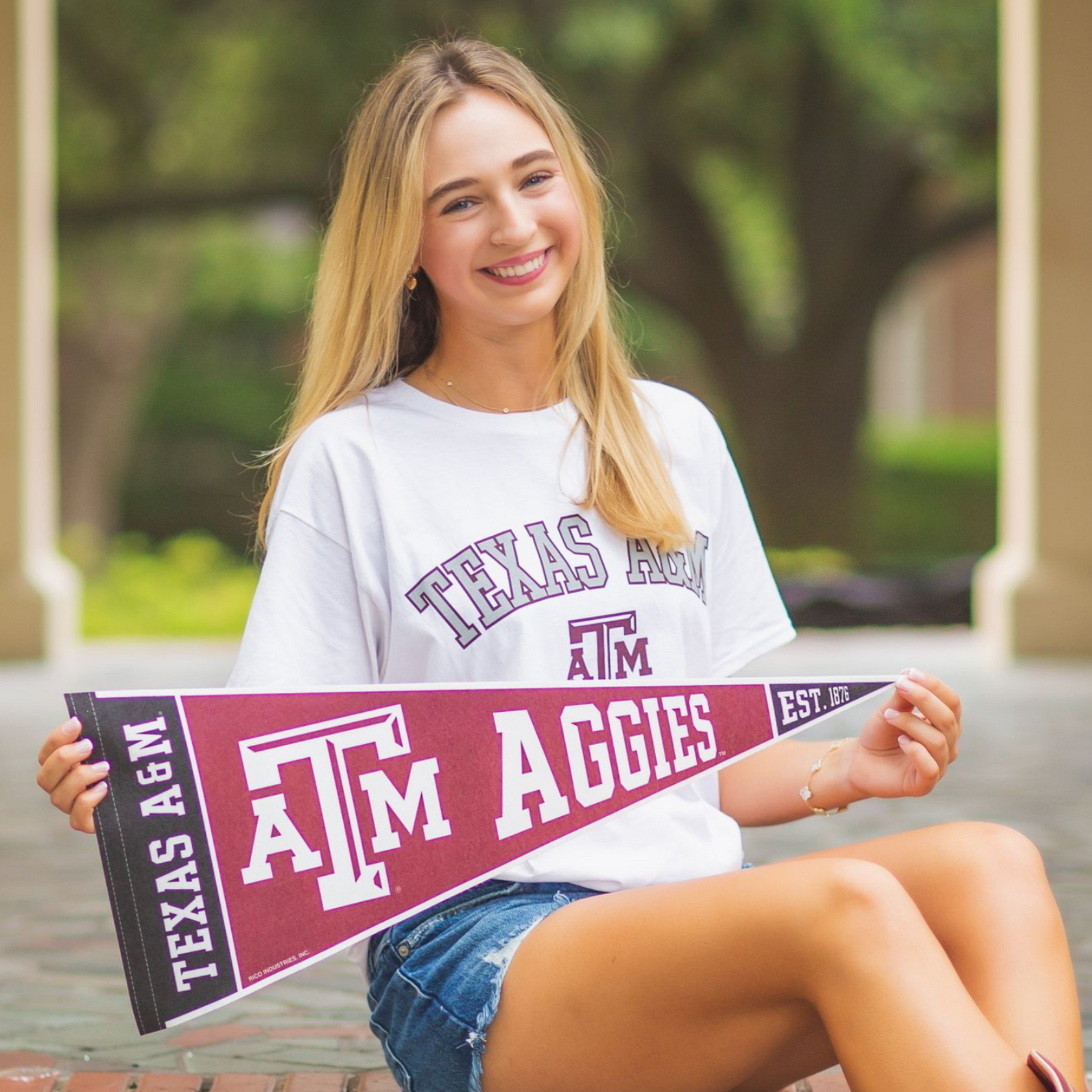 Smiling student holding a maroon Texas A&M pennant while seated outdoors