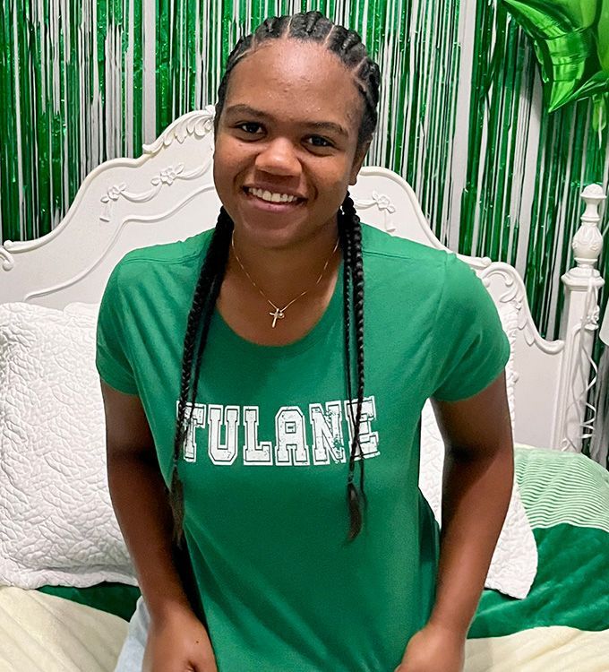 Smiling person in a green Tulane shirt seated on a white bed with green decorations behind them