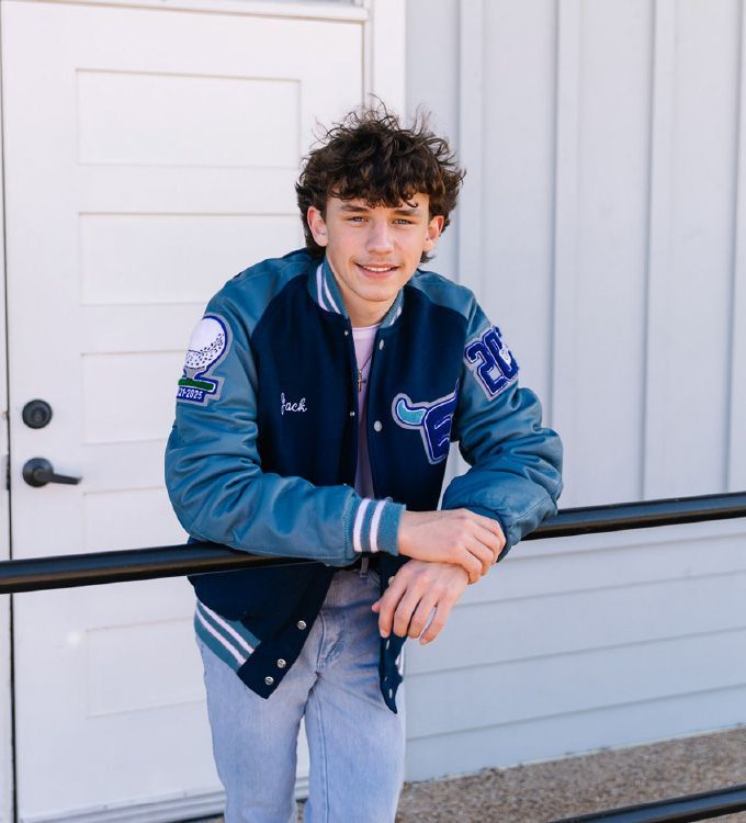 Young person in a blue varsity jacket leaning on a black railing outside a white building
