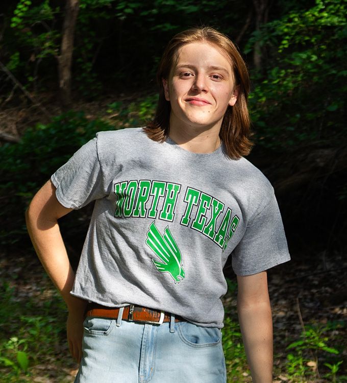 Person in a gray T-shirt and jeans standing outdoors in a wooded area, smiling at the camera.