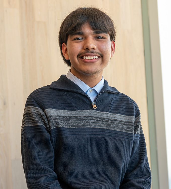 Smiling person in a navy striped sweater standing against a light wood wall