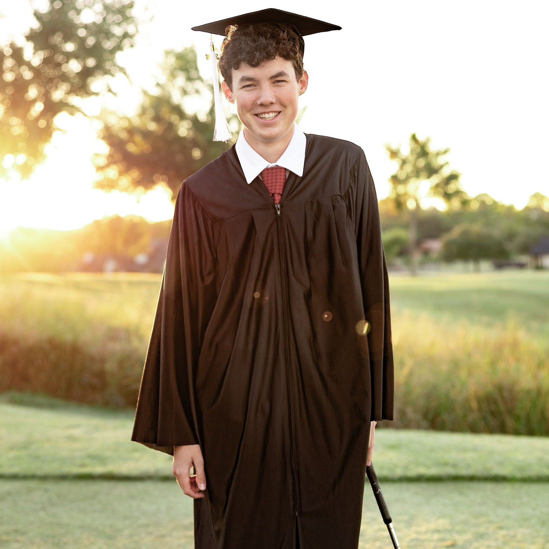 Graduate in brown cap and gown standing outdoors on a sunny lawn, smiling.
