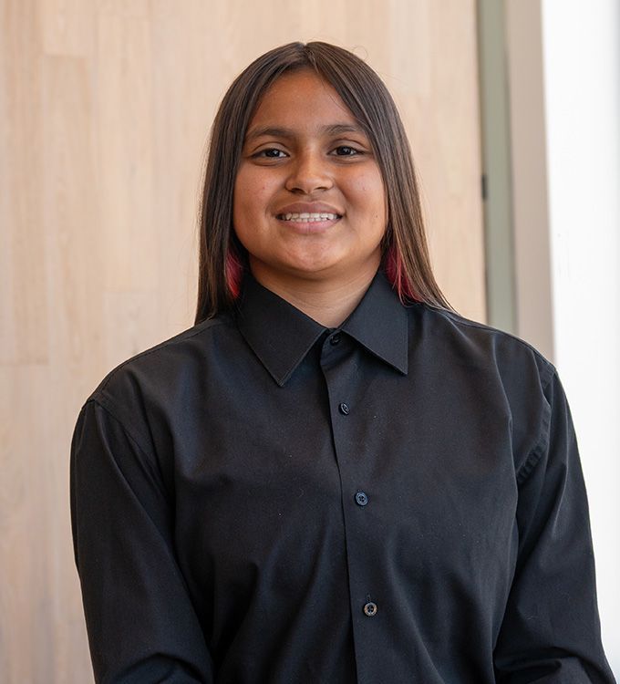 Smiling person in a black button-up shirt standing indoors against a light wood wall.