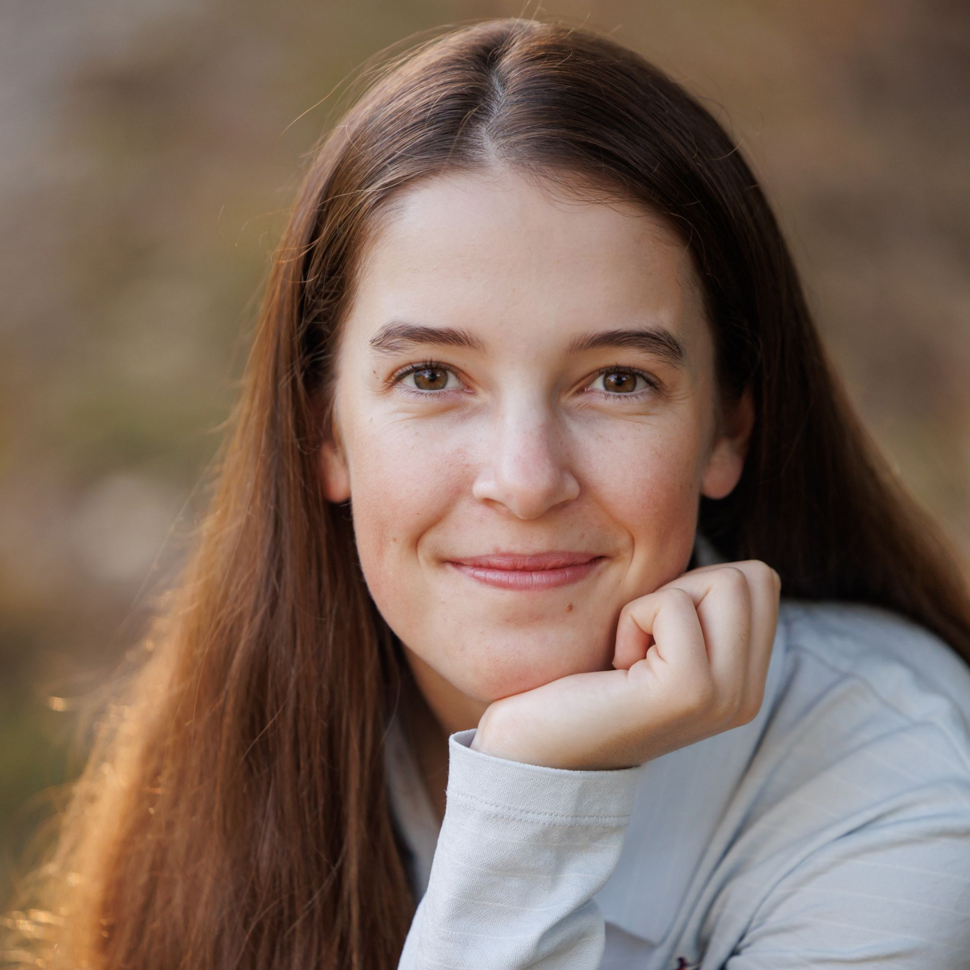 Smiling person with long brown hair resting chin on hand against a blurred outdoor background