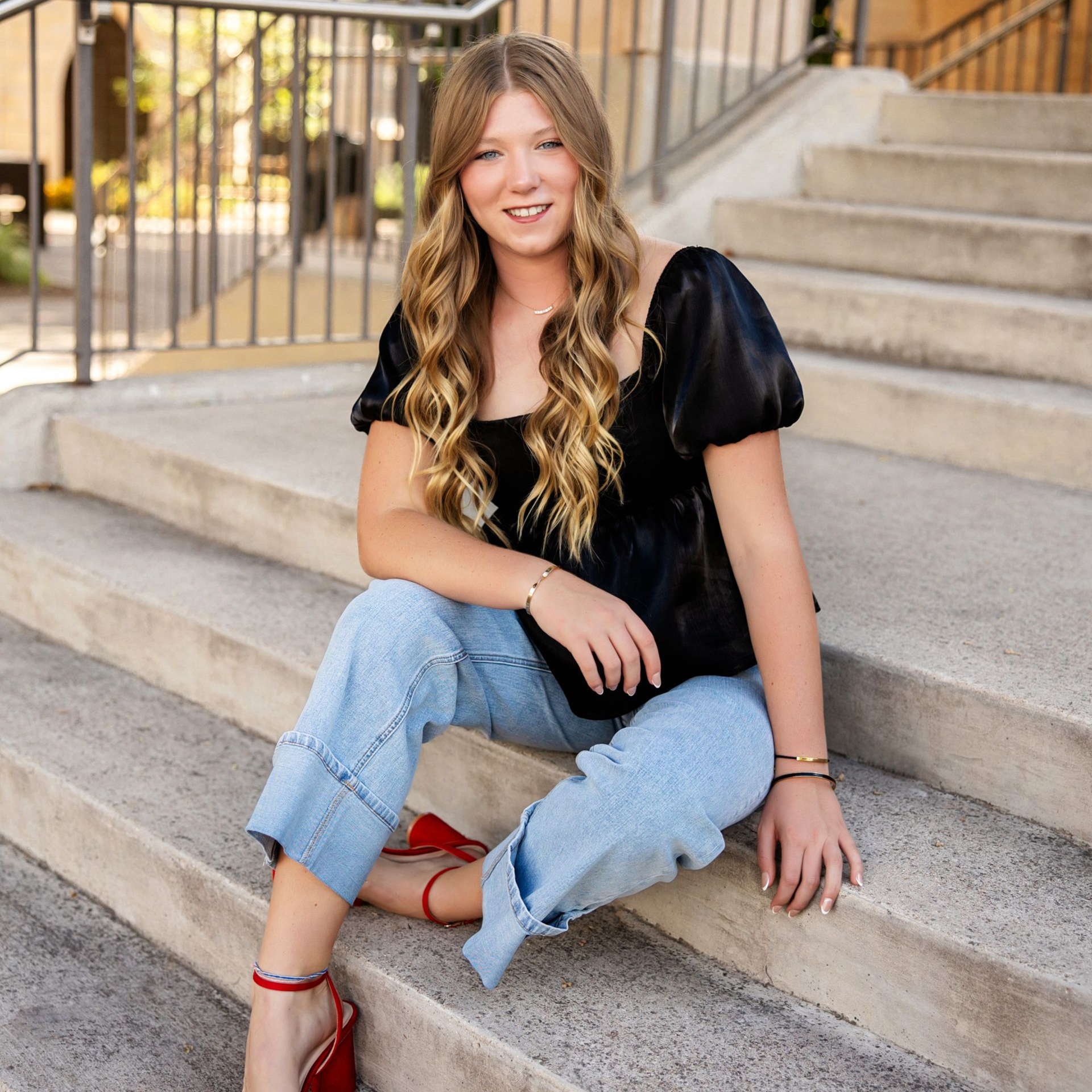Woman sitting on outdoor steps, smiling in a black blouse, light jeans, and red heels.