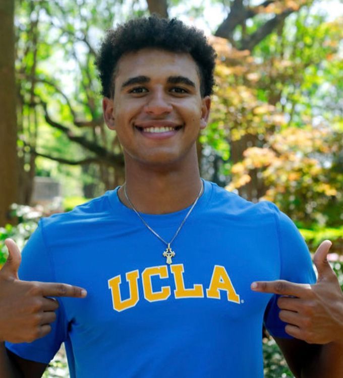 Smiling person in blue UCLA shirt pointing at the UCLA logo outdoors