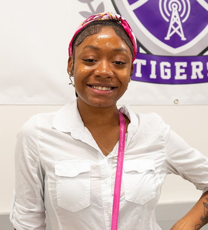 Smiling woman in white shirt and pink headband standing before a purple Tigers banner