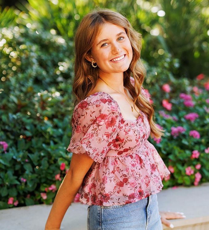 Smiling woman in a pink floral top and jeans standing outdoors in front of green foliage and flowers