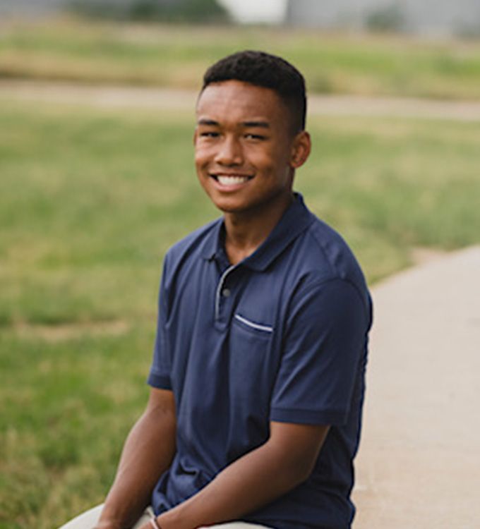 Smiling person in a navy polo shirt sitting outdoors on a sidewalk beside grass.
