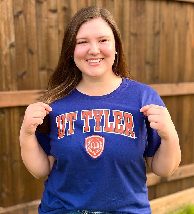 Smiling person in a blue UT Tyler T-shirt standing in front of a wooden fence.