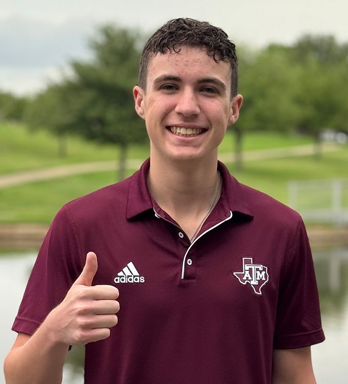 Smiling person in maroon polo giving a thumbs-up outdoors on a green campus lawn