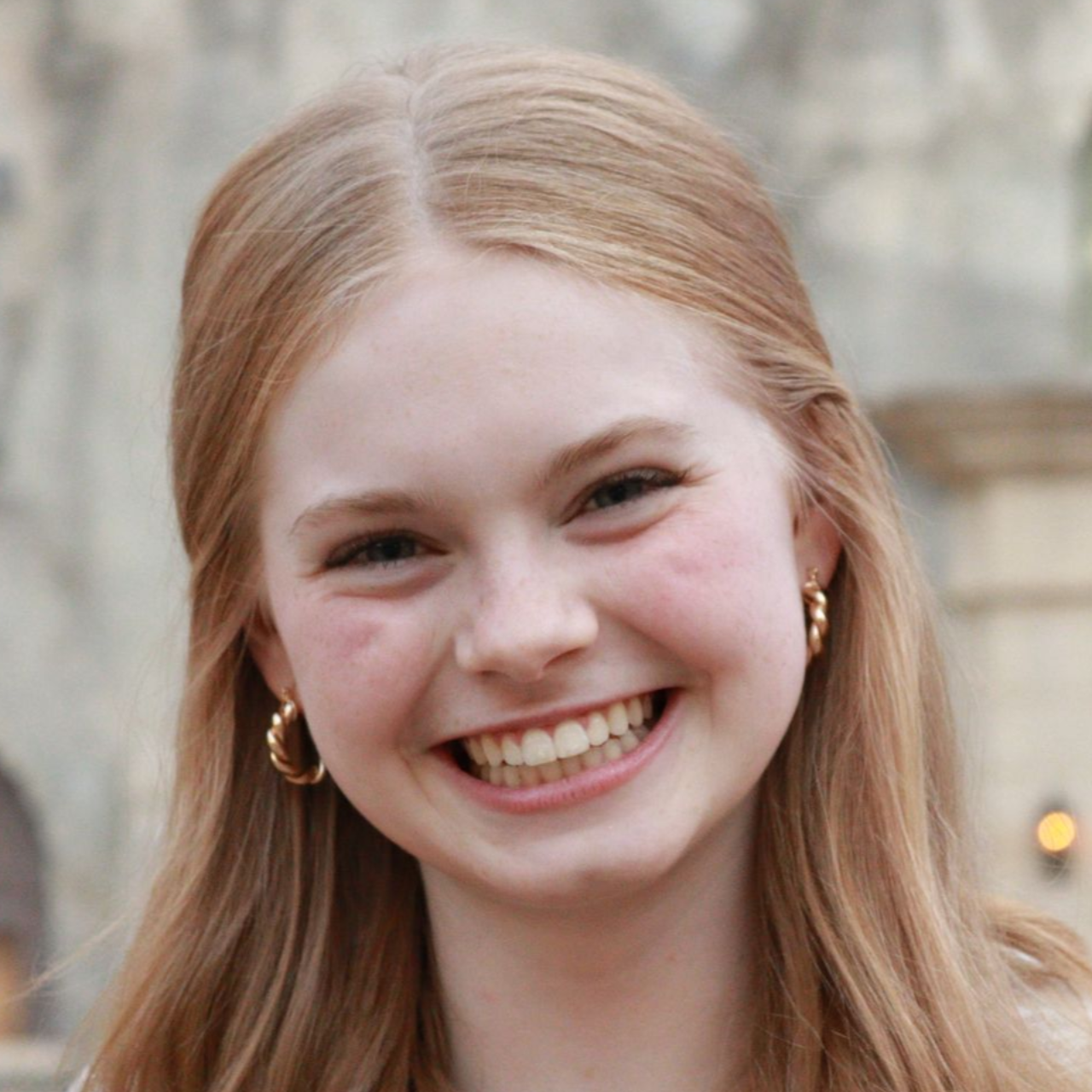 Smiling person with blonde hair and gold hoop earrings, outdoors against a blurred stone background