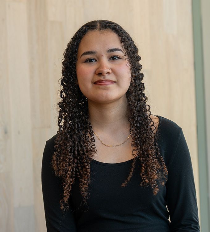 Portrait of a woman with curly hair in a black top, standing against a light wood wall