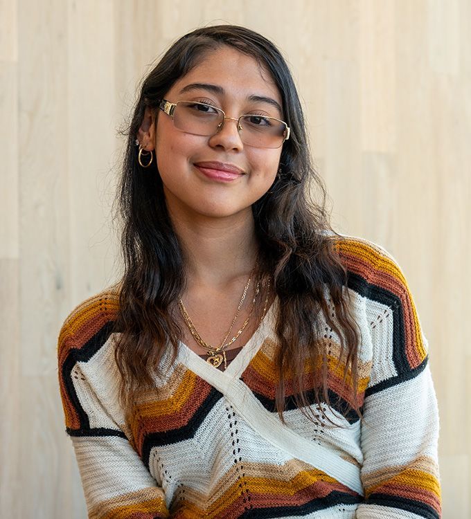 Smiling person with glasses in a striped sweater, seated against a light wood wall.