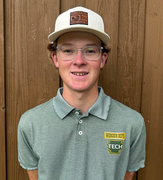 Person wearing a white cap and glasses, standing against a wooden wall in a green polo shirt.