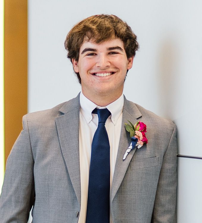 Smiling person in a gray suit with a navy tie and pink boutonniere against a light wall.