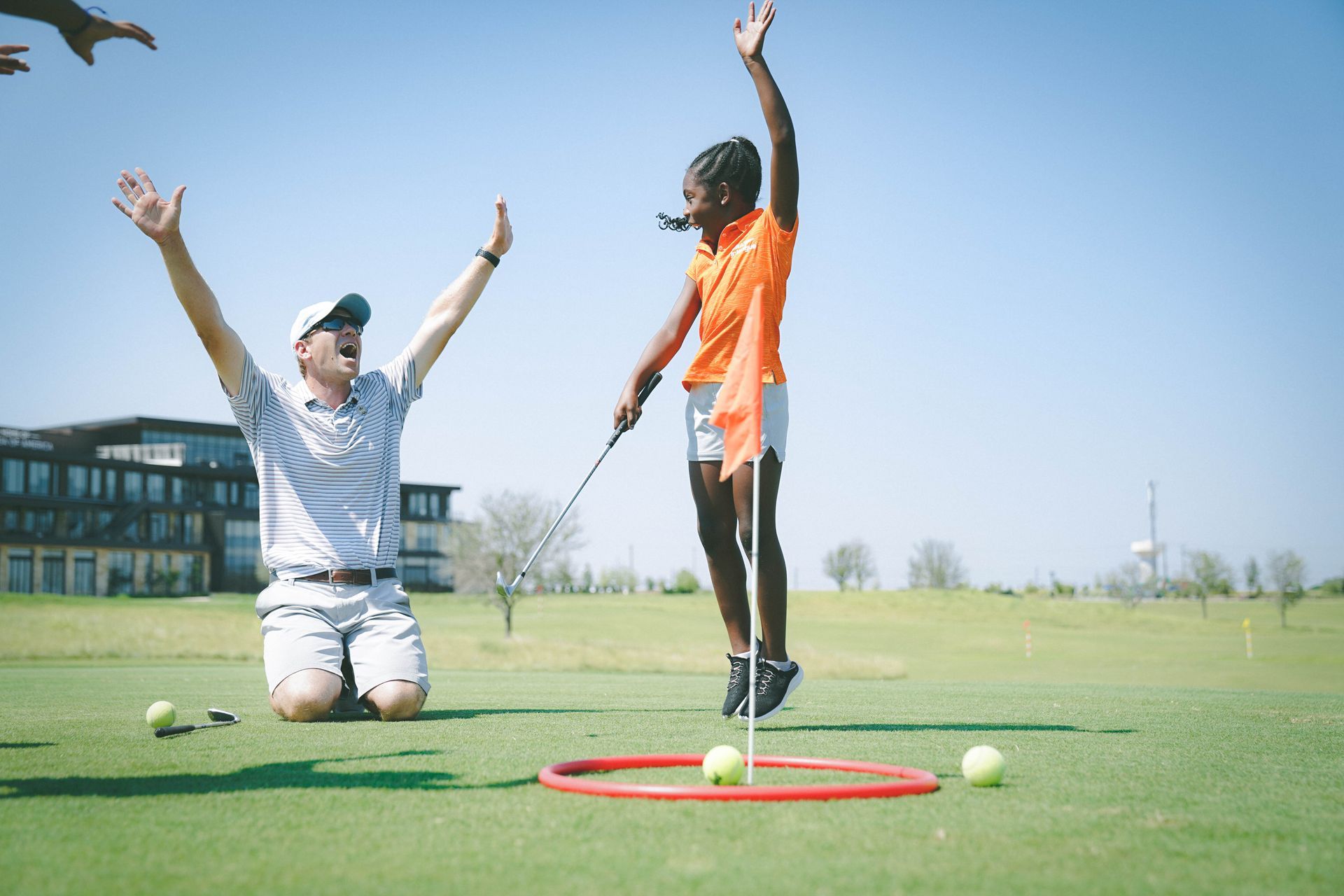 Two people on a golf course celebrating with arms raised, one kneeling and one standing by a red hoop