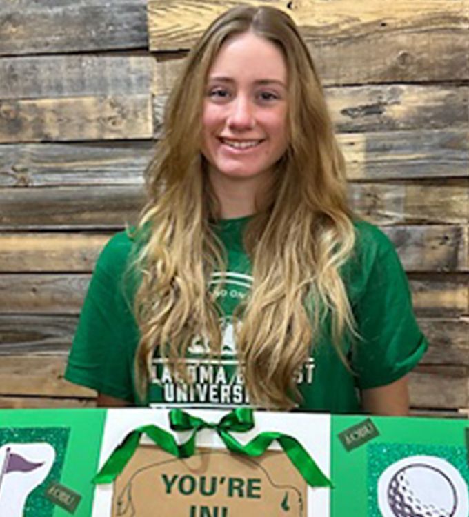 Smiling student in green shirt behind a green-and-white university sign against a wood wall