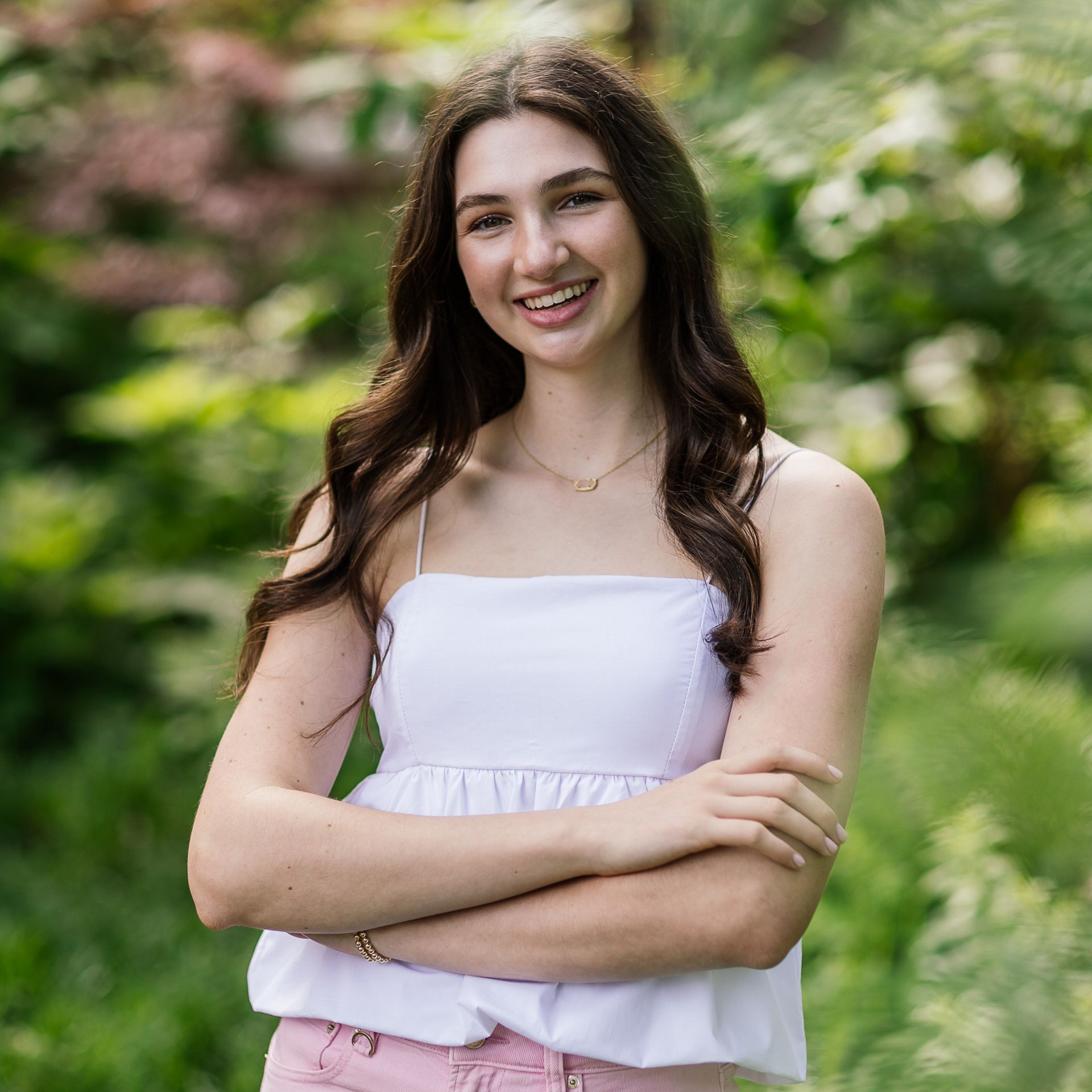 Smiling woman in a white top stands outdoors with arms crossed in a green garden.