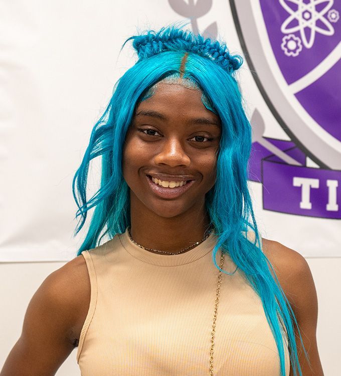 Smiling person with bright blue hair and beige top standing before a purple logo backdrop