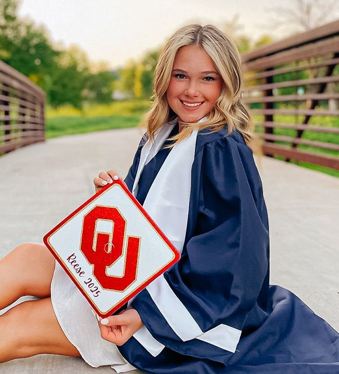 Smiling student in navy graduation gown holding an OU sign outdoors on a bridge-like walkway