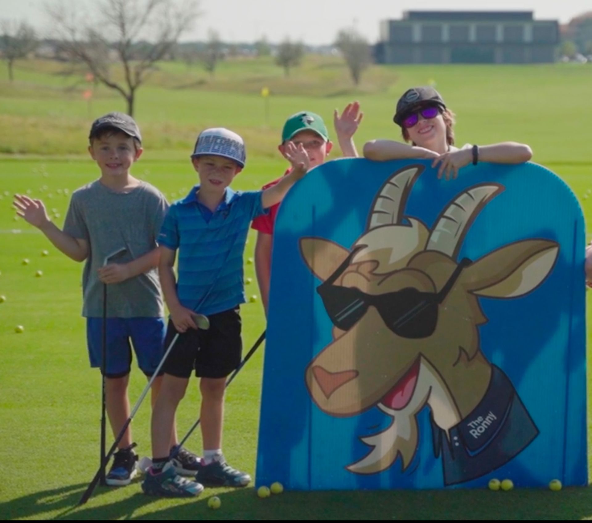 Four kids on a golf course pose beside a cartoon kangaroo sign with sunglasses.