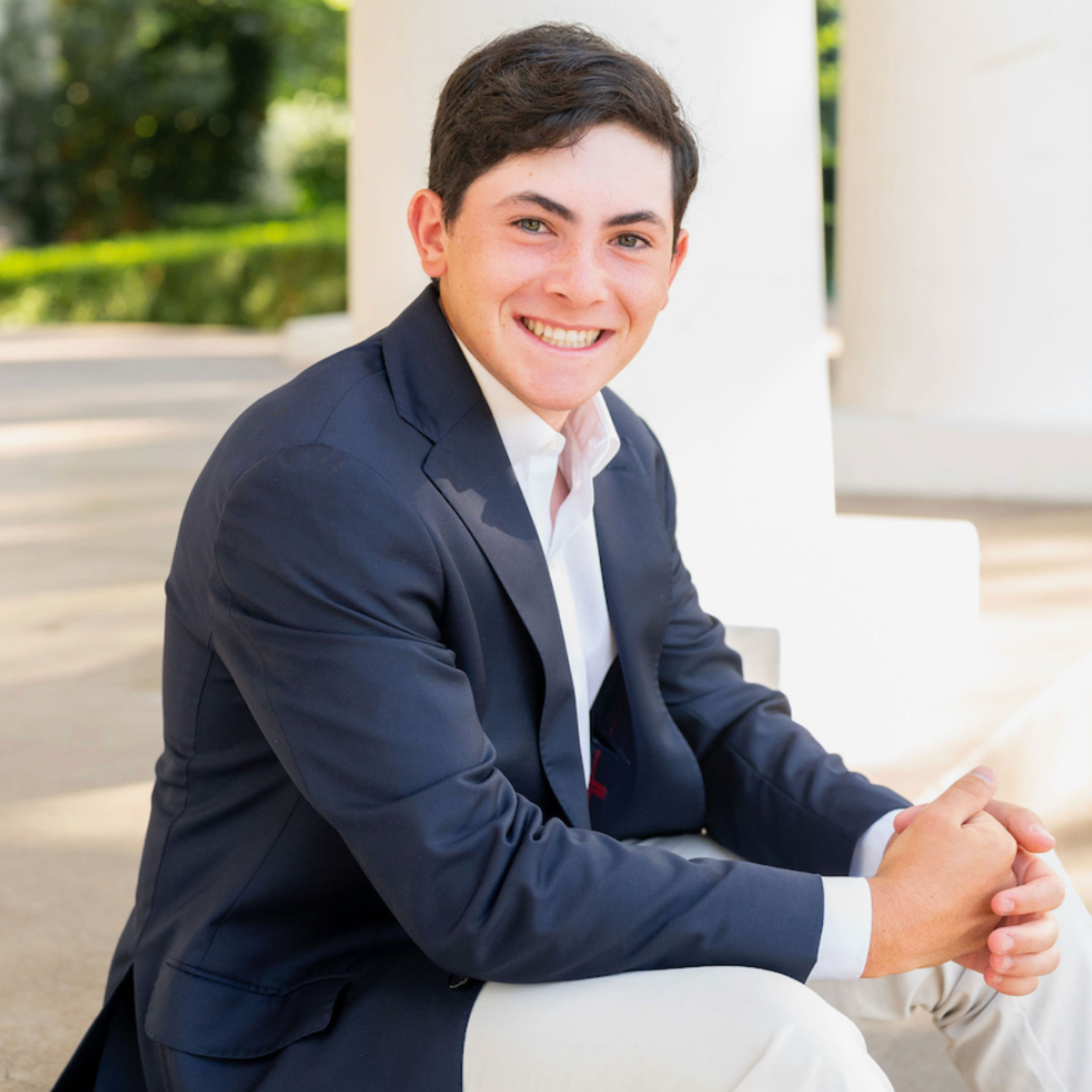 Smiling person in a navy blazer seated outdoors near a white column