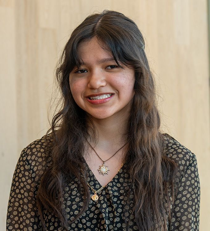 Smiling person with long dark hair in a patterned black top against a light wood wall