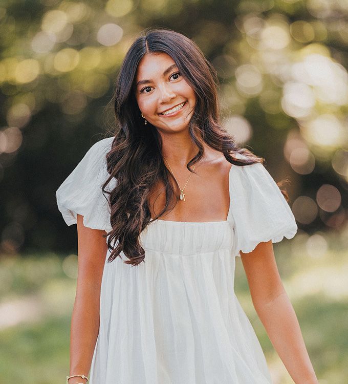 Smiling woman in a white dress standing outdoors in soft sunlight with blurred greenery behind her